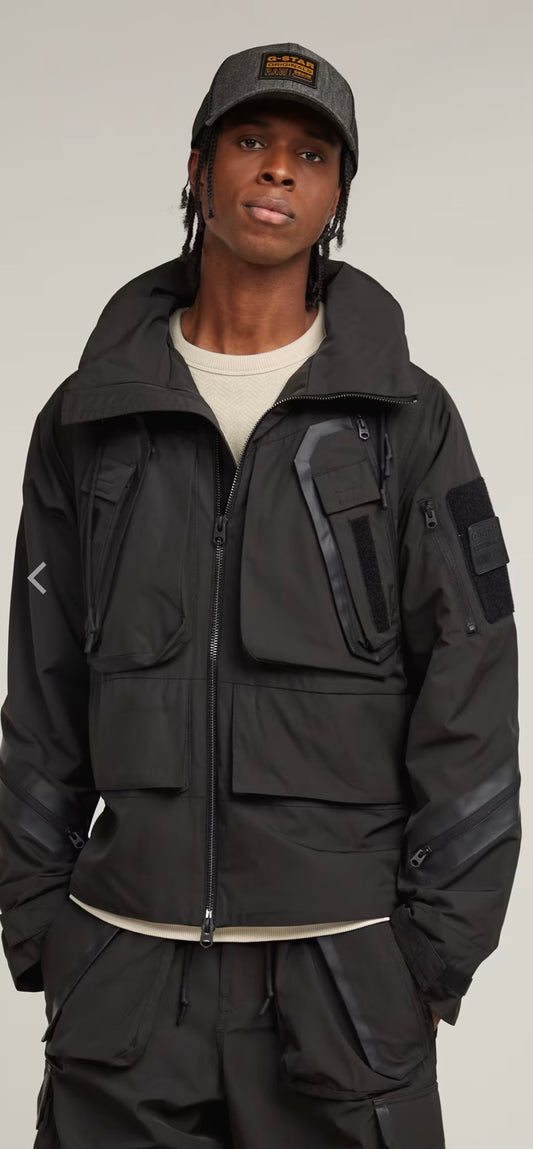 Person wearing a black tactical jacket and cap on a gray background